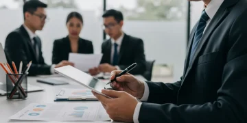 Profissional de auditoria interna revisando documentos e dados em um tablet, com equipe ao fundo.